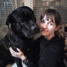 Junge Frau (Rebecca, Inhaberin der Website) mit dunkler Kleidung umarmt einen großen schwarzen Hund.